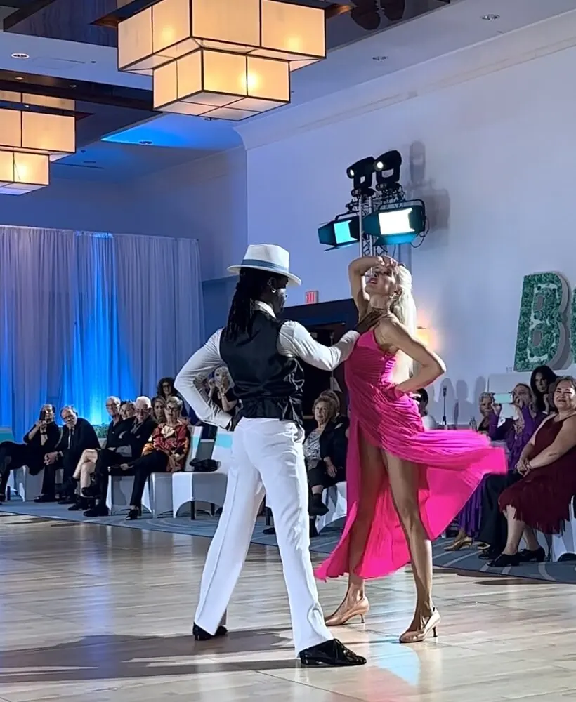 Ballroom couple performing for a seated audience, she in a pink dress, he in a white fedora